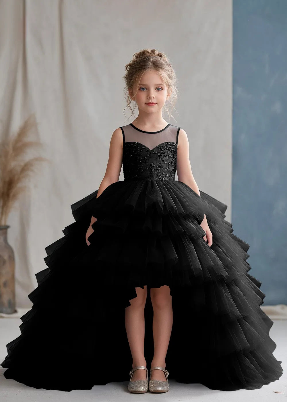 Front view of a child wearing a black flower girl dress with a sheer illusion neckline over a beaded bodice and a high-low tulle skirt with broad tiered ruffle layers.