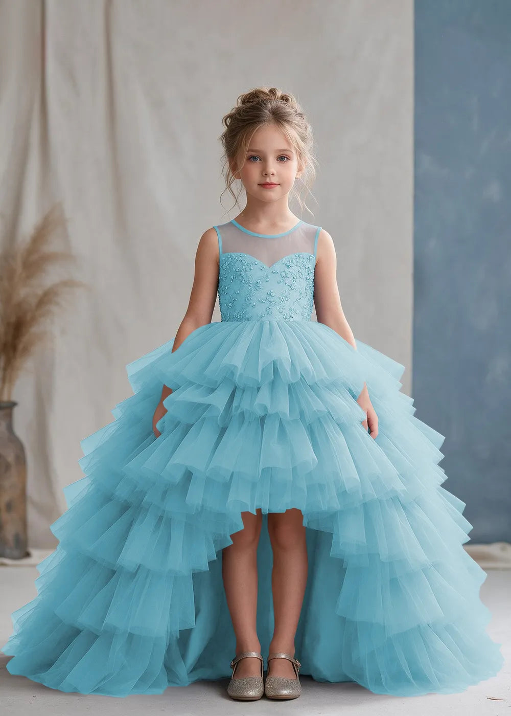 Front view of a child wearing a blue flower girl dress with a sheer illusion neckline over a beaded bodice and a high-low tulle skirt with broad tiered ruffle layers.