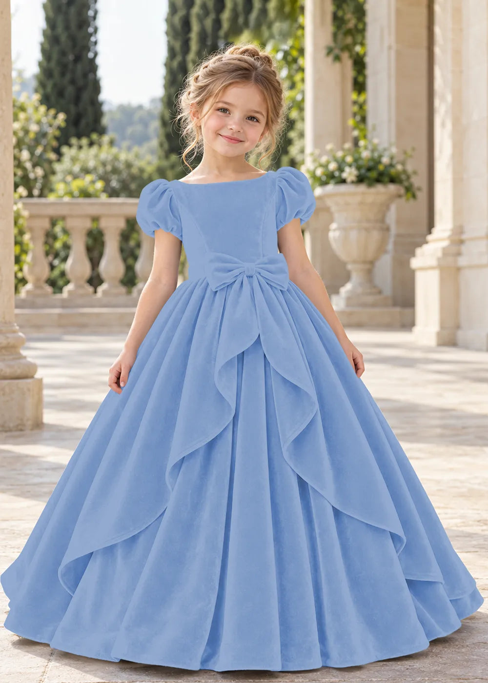 Front view of a child wearing a blue flower girl dress with a square neckline, short puff sleeves and a large velvet bow above a full tiered skirt.