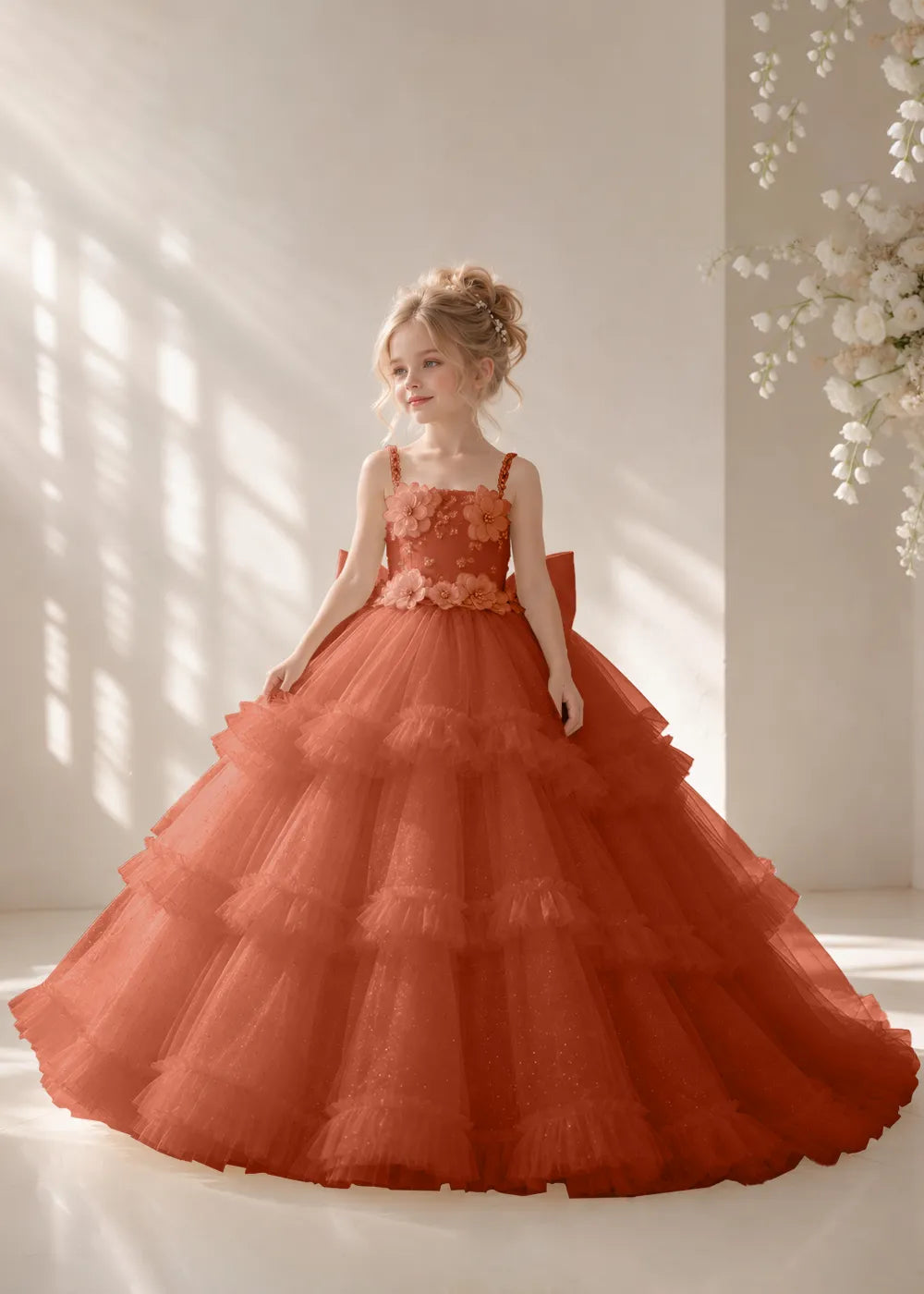 Front view of a child wearing a burnt orange flower girl dress with beaded spaghetti straps, a bodice covered in three-dimensional fabric flowers and a multi-tiered tulle skirt.