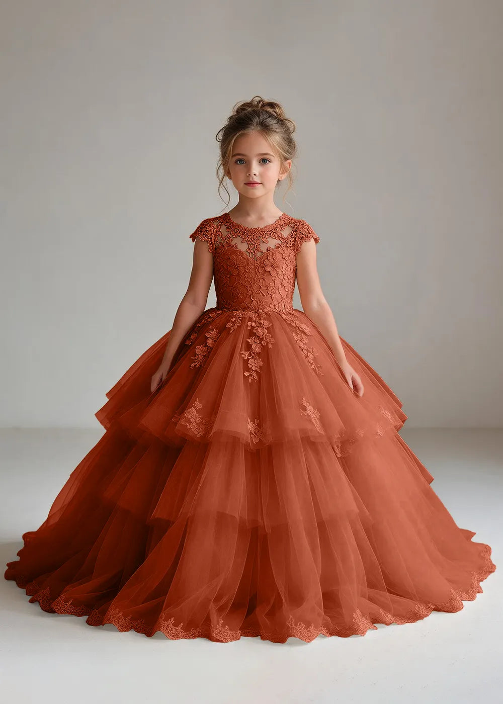Front view of a child wearing a burnt orange flower girl dress with a sheer lace illusion neckline, cap sleeves and a tiered cake-style tulle skirt with a lace-trimmed hem.
