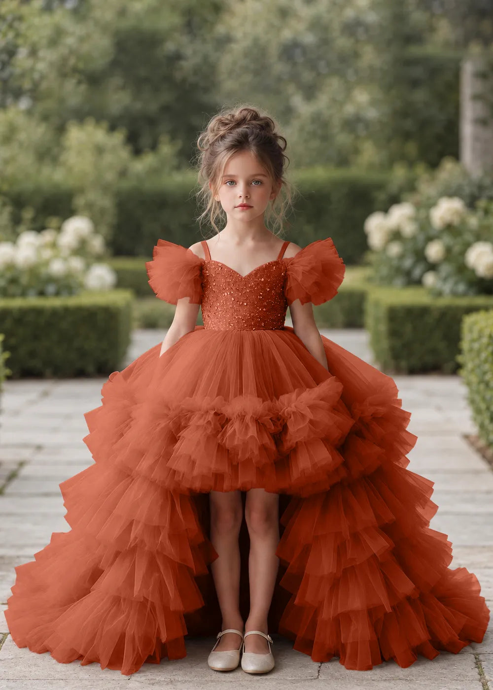 Front view of a child wearing a burnt orange flower girl dress with a V-neckline, a sequin-covered bodice and a tiered high-low tulle skirt with off-shoulder sleeve ruffles.