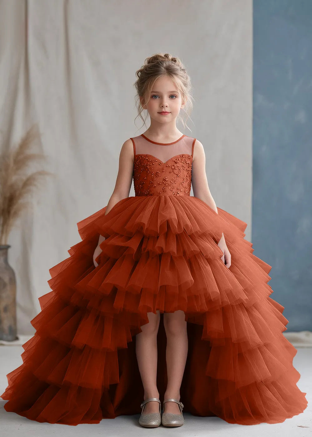 Front view of a child wearing a burnt orange flower girl dress with a sheer illusion neckline over a beaded bodice and a high-low tulle skirt with broad tiered ruffle layers.