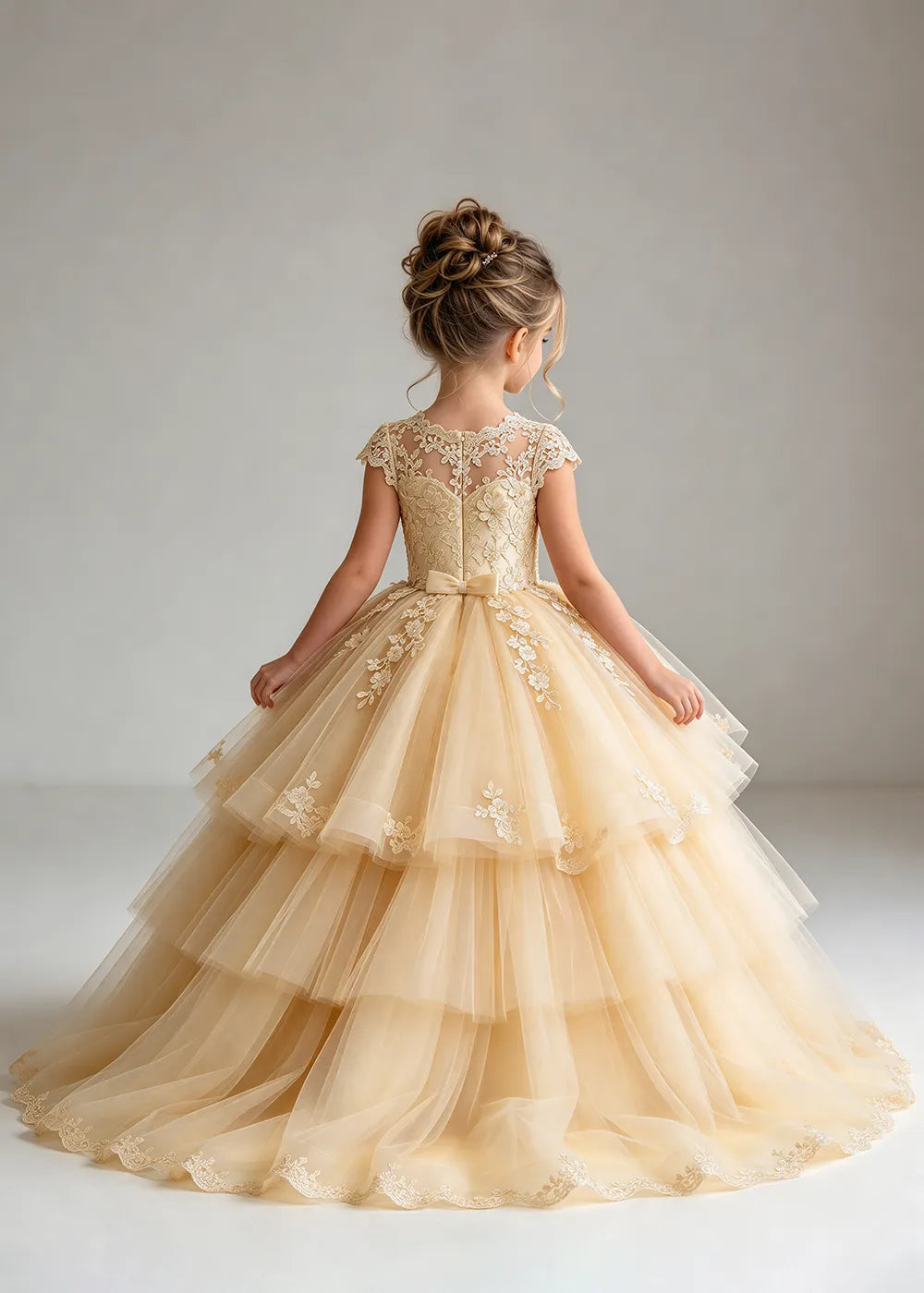 The rear view of a child wearing a Champagne flower girl dress with a sheer lace illusion neckline, cap sleeves and a tiered cake-style tulle skirt with a lace-trimmed hem.