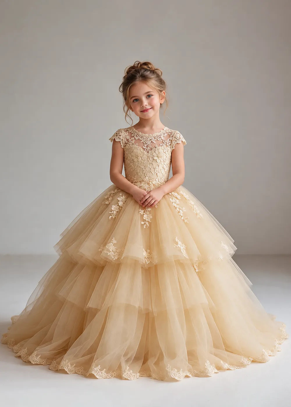 The front view of a child wearing a Champagne flower girl dress with a sheer lace illusion neckline, cap sleeves and a tiered cake-style tulle skirt with a lace-trimmed hem.