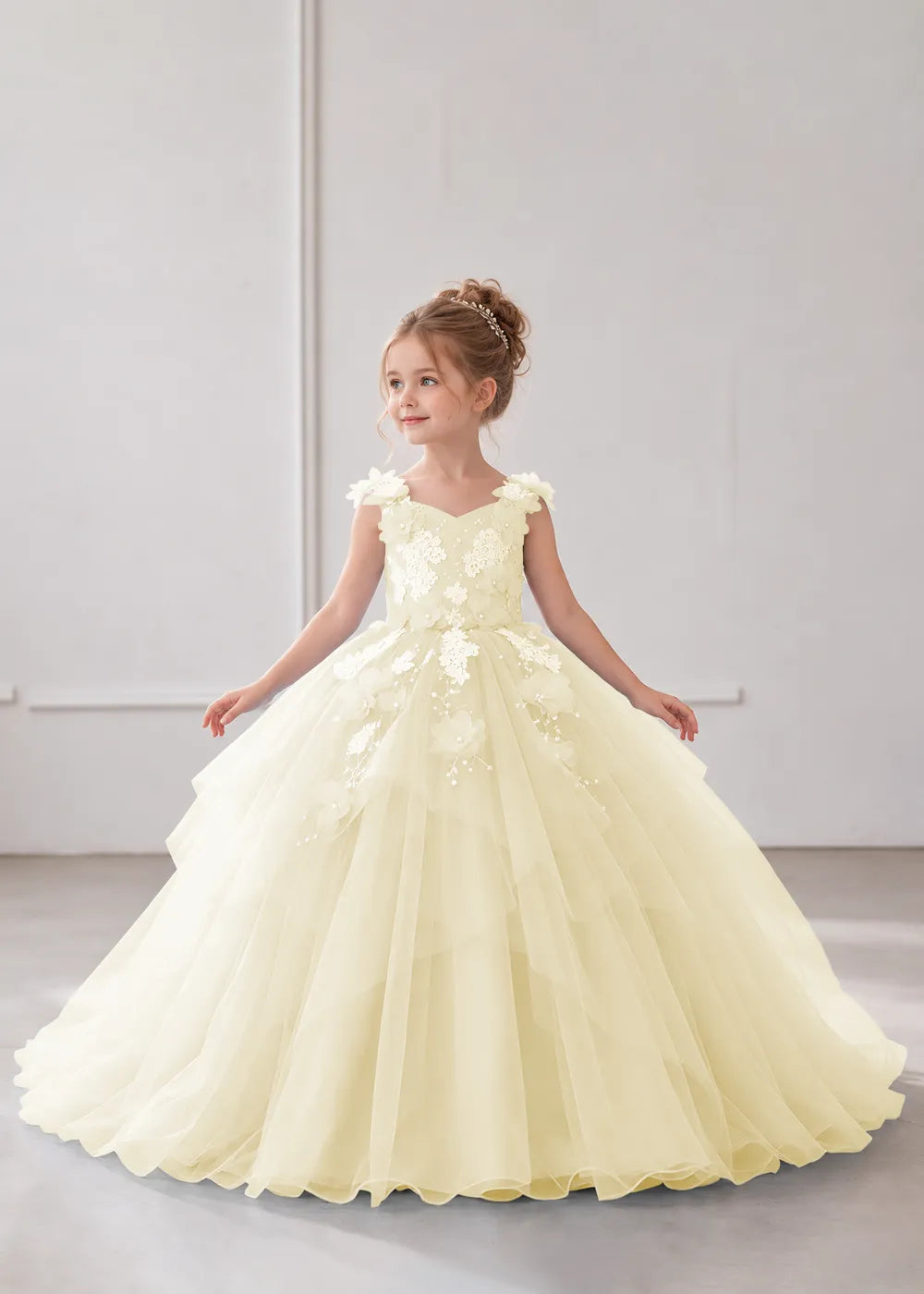 Front view of a child wearing a champagne flower girl dress with a V-neckline, floral shoulder appliques and lace and flower decoration across the bodice and hips.