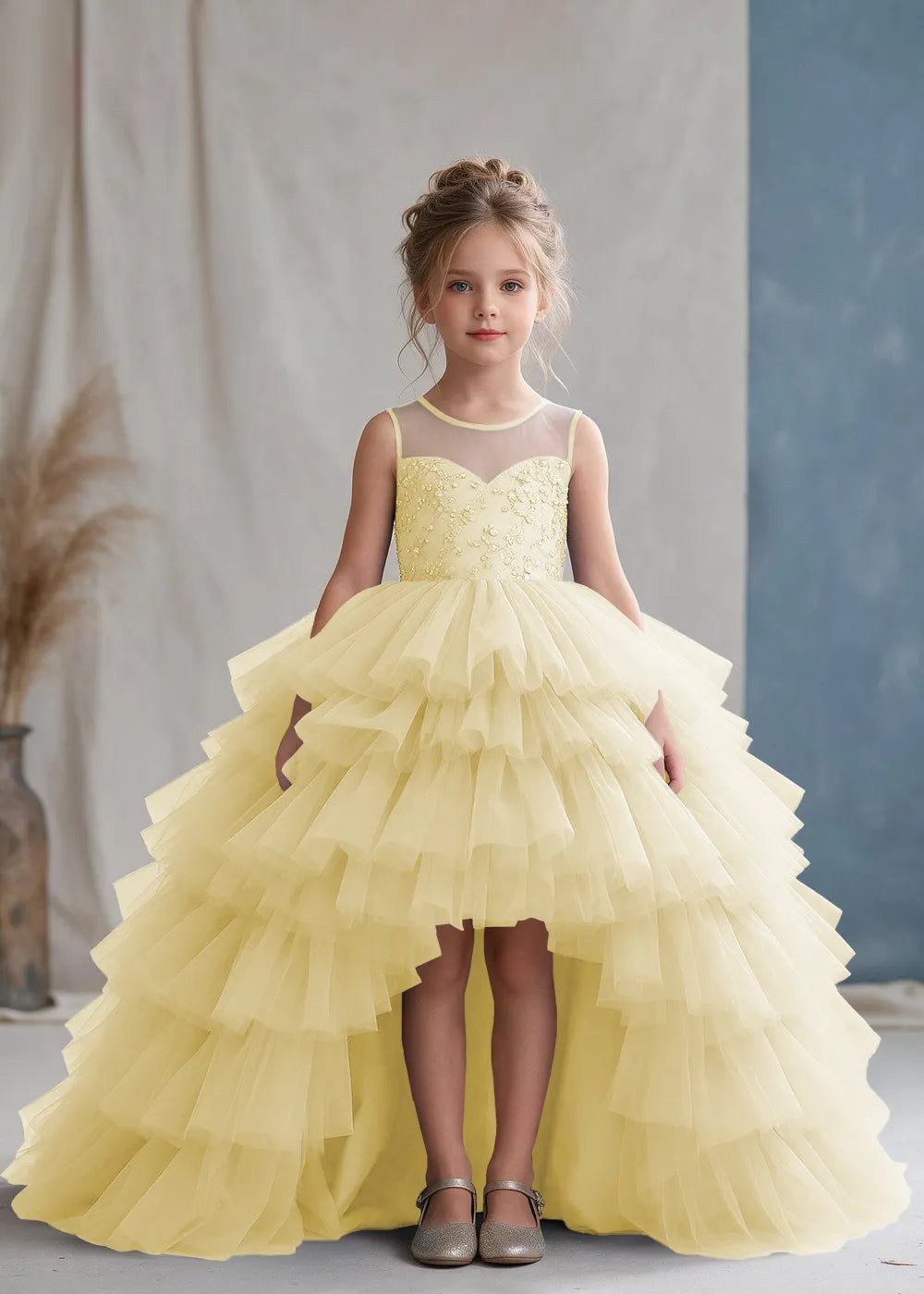 Front view of a child wearing a champagne flower girl dress with a sheer illusion neckline over a beaded bodice and a high-low tulle skirt with broad tiered ruffle layers.