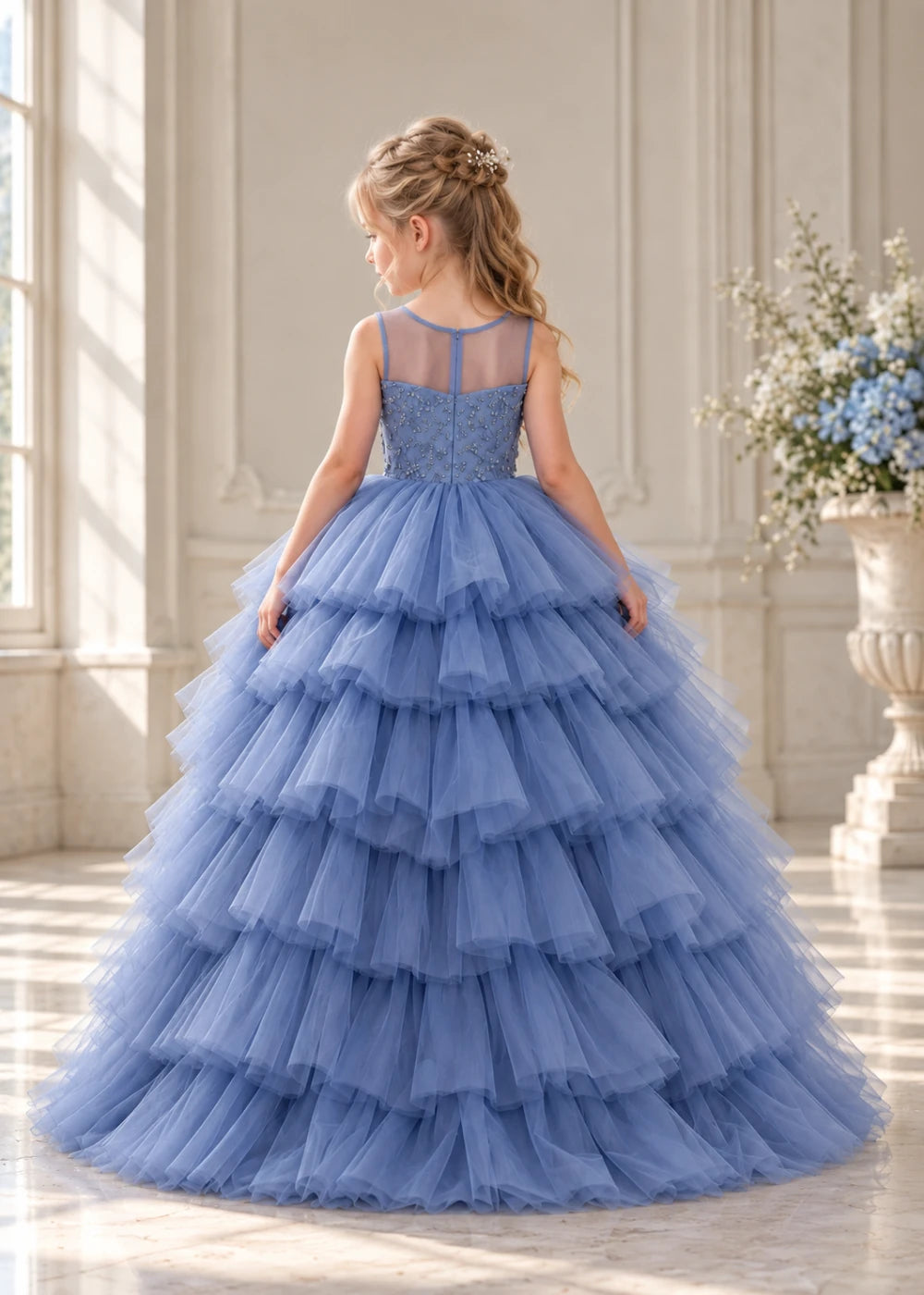 Rear view of a child wearing a dusty blue flower girl dress with a sheer illusion neckline over a beaded bodice and a high-low tulle skirt with broad tiered ruffle layers.