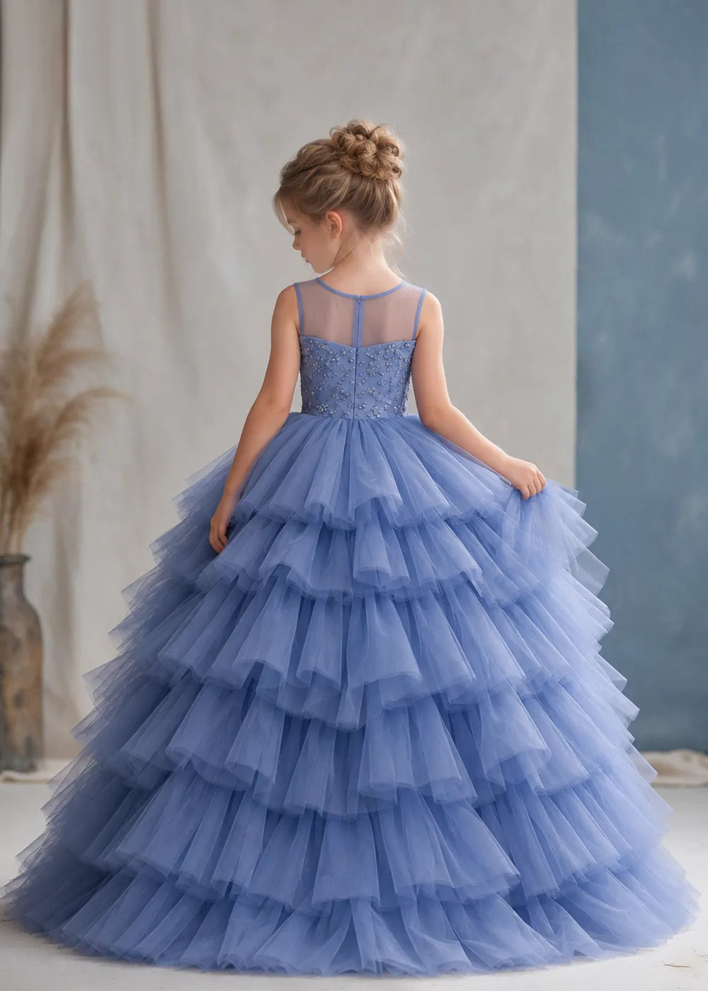 The rear view of a child wearing a Dusty Blue flower girl dress with a sheer illusion neckline over a beaded bodice and a high-low tulle skirt with broad tiered ruffle layers.
