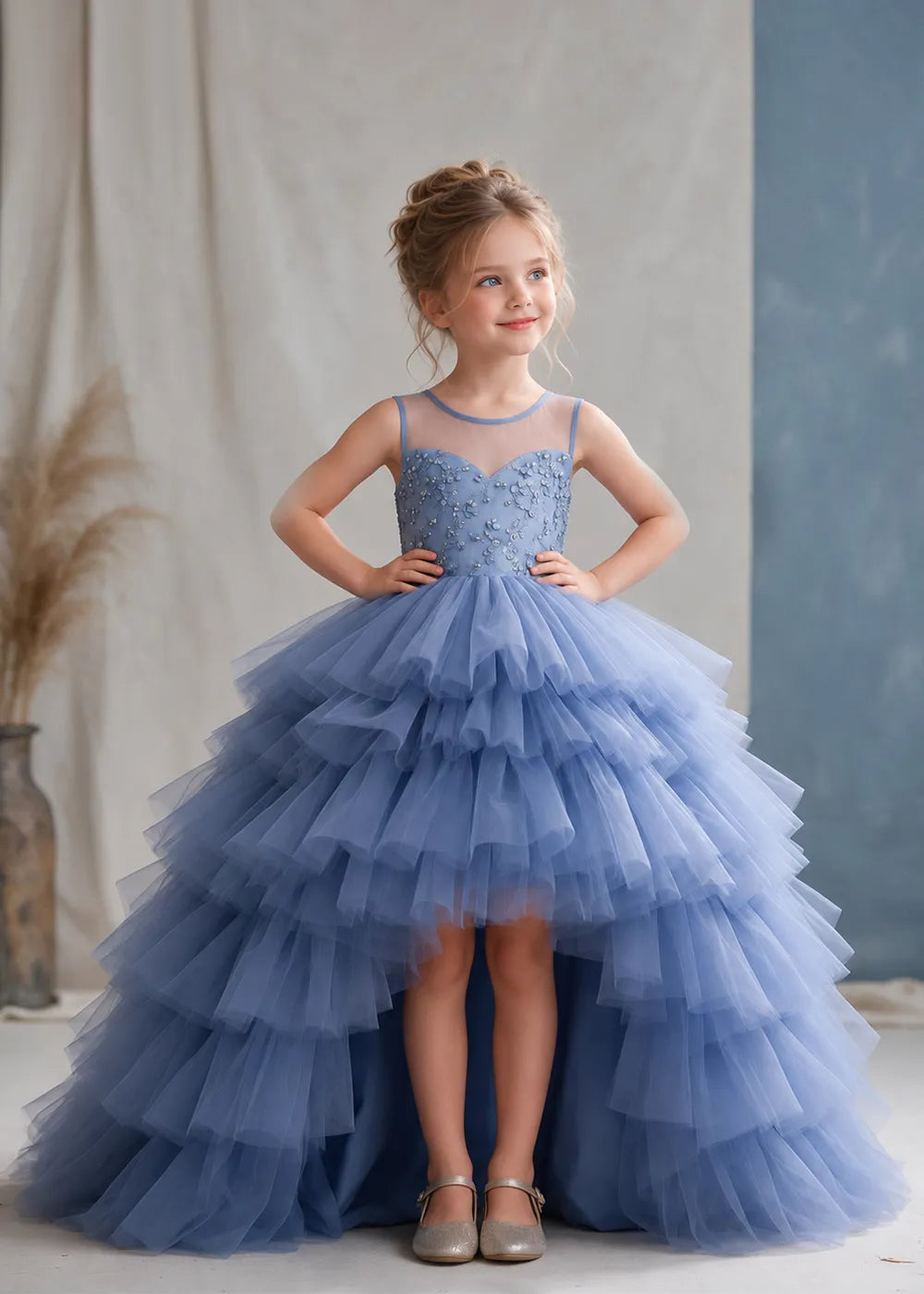 The three-quarter view of a child wearing a Dusty Blue flower girl dress with a sheer illusion neckline over a beaded bodice and a high-low tulle skirt with broad tiered ruffle layers.