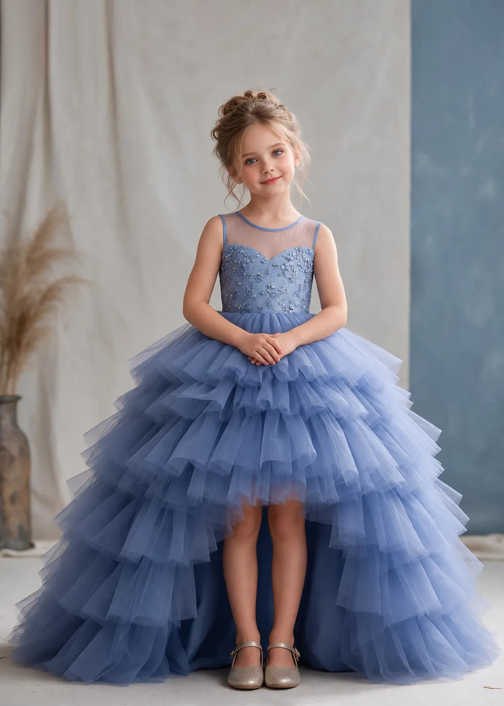The front view of a child wearing a Dusty Blue flower girl dress with a sheer illusion neckline over a beaded bodice and a high-low tulle skirt with broad tiered ruffle layers.