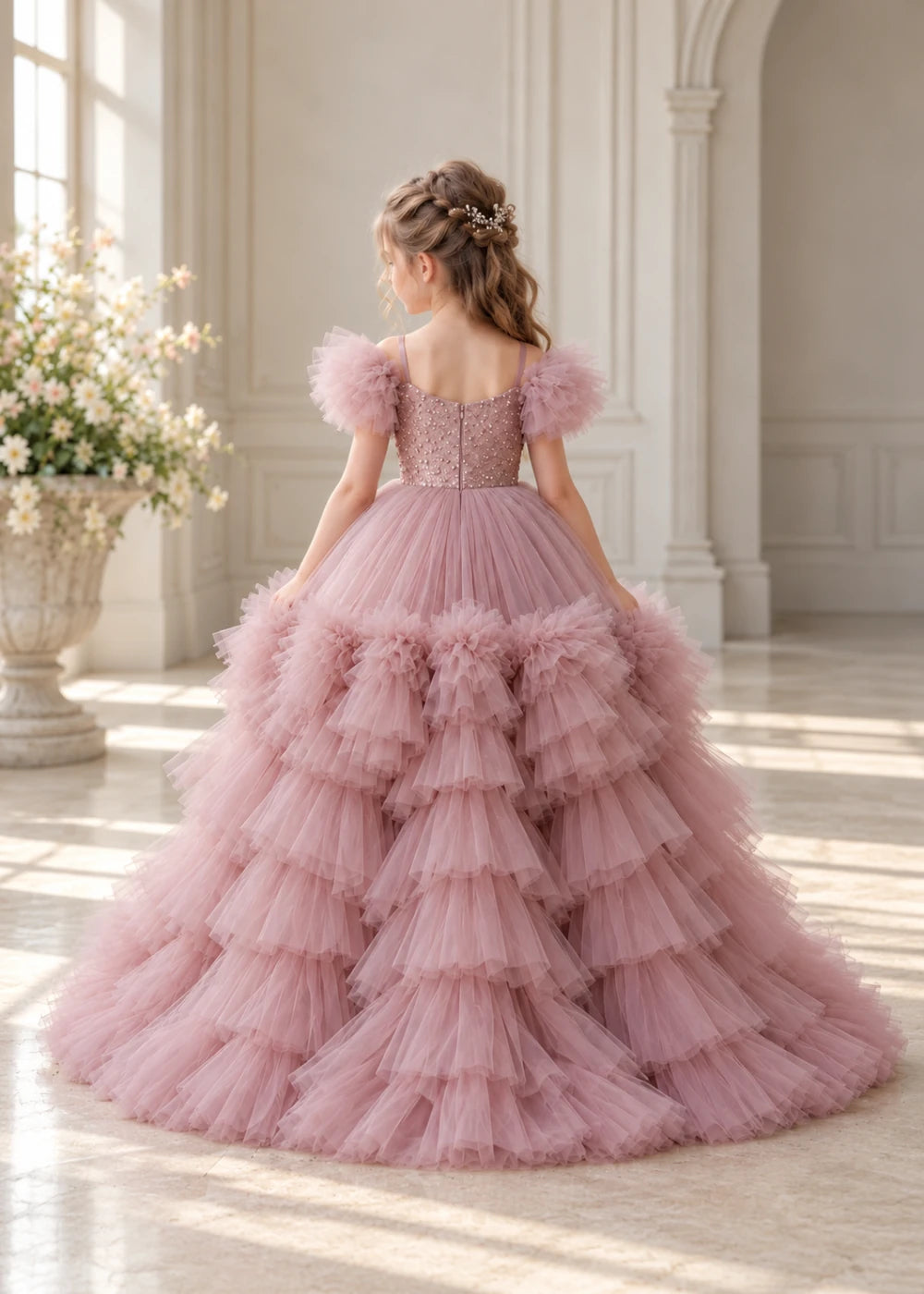 Rear view of a child wearing a dusty rose flower girl dress with a V-neckline, a sequin-covered bodice and a tiered high-low tulle skirt with off-shoulder sleeve ruffles.