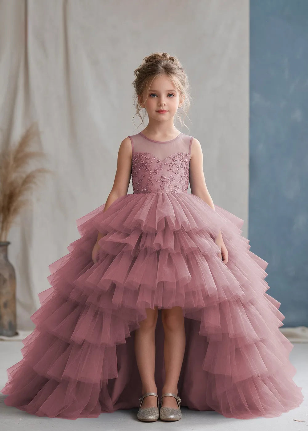 Front view of a child wearing a dusty rose flower girl dress with a sheer illusion neckline over a beaded bodice and a high-low tulle skirt with broad tiered ruffle layers.