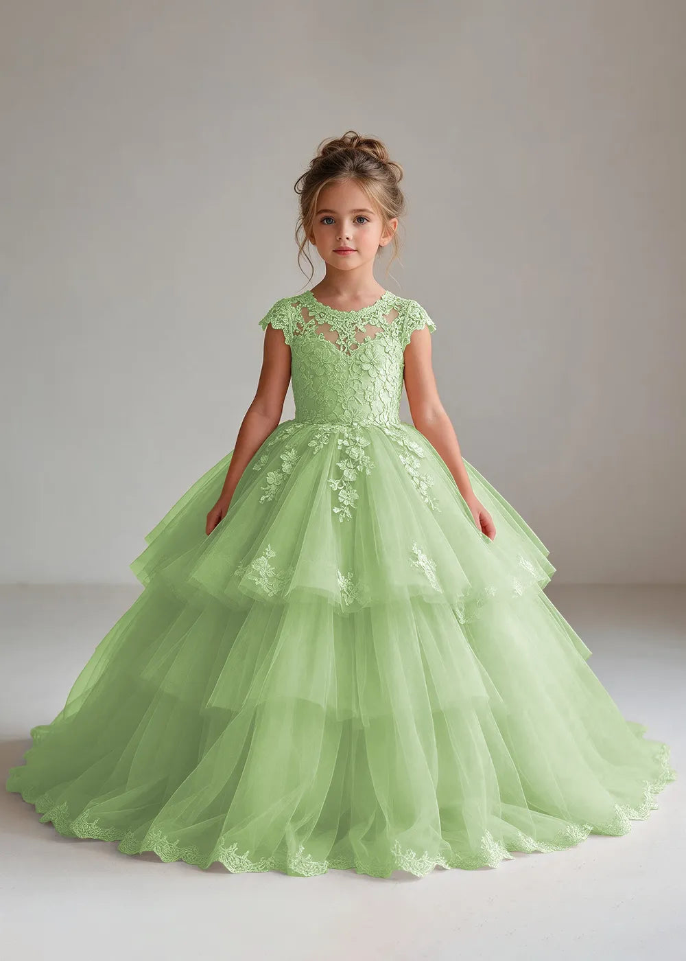 Front view of a child wearing a dusty sage flower girl dress with a sheer lace illusion neckline, cap sleeves and a tiered cake-style tulle skirt with a lace-trimmed hem.
