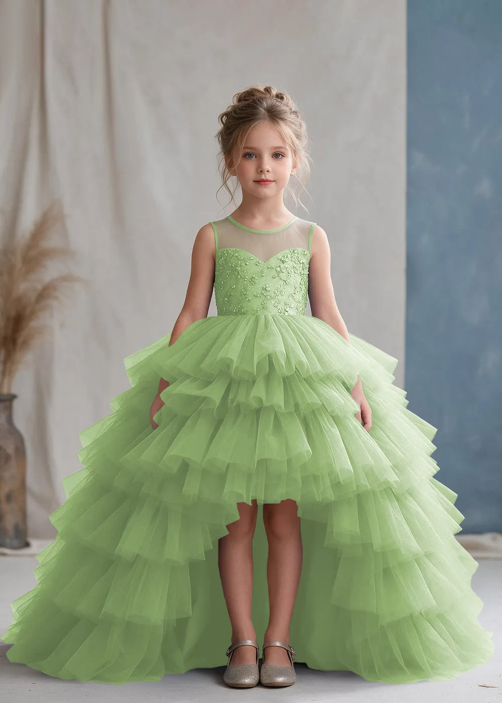 Front view of a child wearing a dusty sage flower girl dress with a sheer illusion neckline over a beaded bodice and a high-low tulle skirt with broad tiered ruffle layers.
