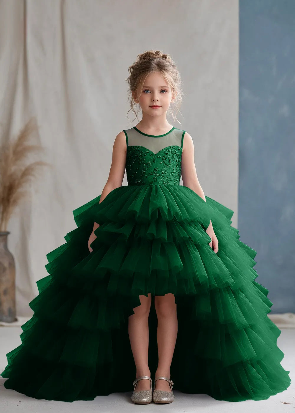 Front view of a child wearing a emerald green flower girl dress with a sheer illusion neckline over a beaded bodice and a high-low tulle skirt with broad tiered ruffle layers.