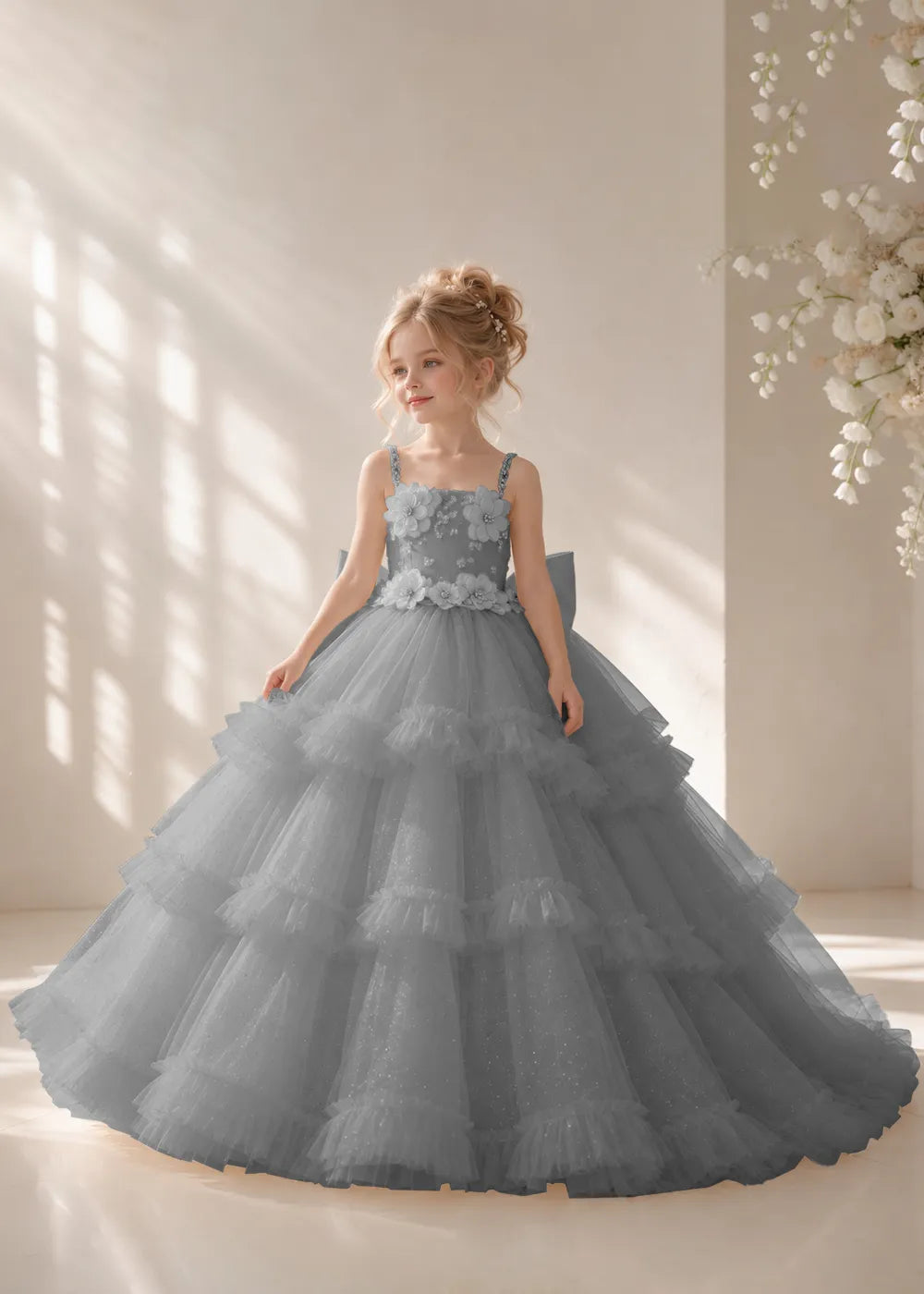 Front view of a child wearing a grey flower girl dress with beaded spaghetti straps, a bodice covered in three-dimensional fabric flowers and a multi-tiered tulle skirt.