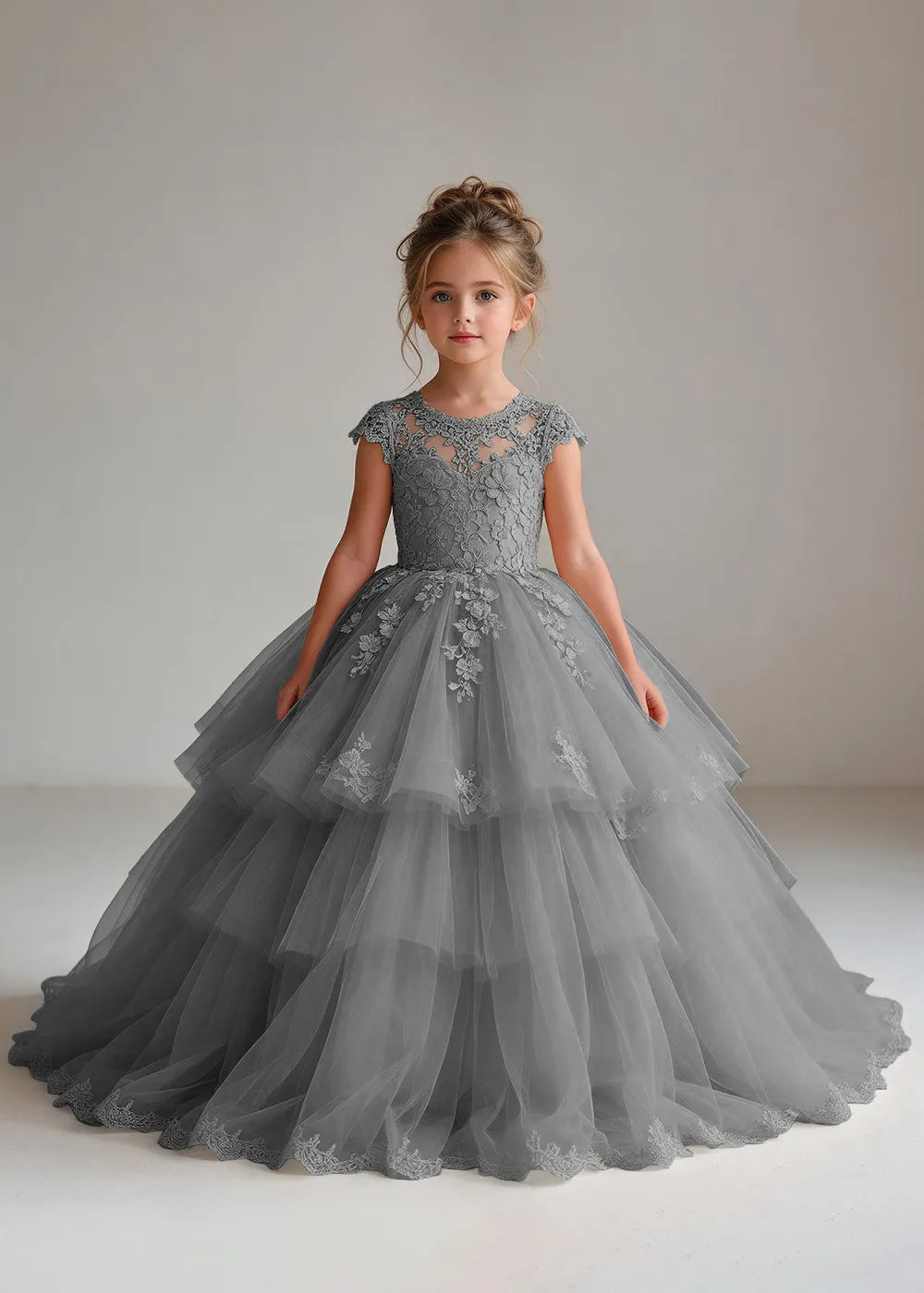 Front view of a child wearing a grey flower girl dress with a sheer lace illusion neckline, cap sleeves and a tiered cake-style tulle skirt with a lace-trimmed hem.