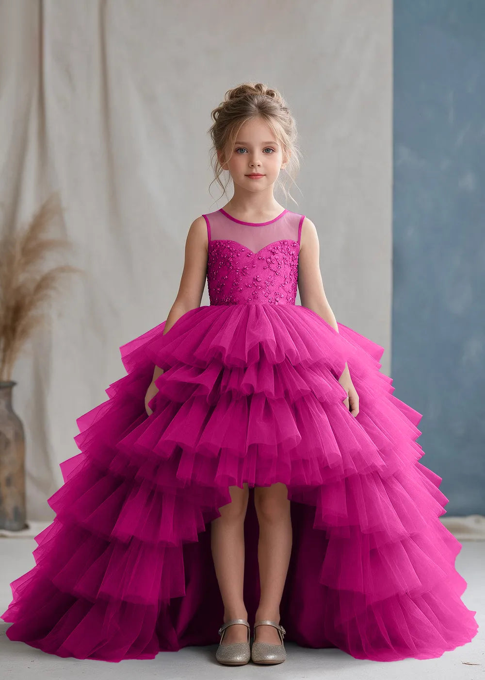 Front view of a child wearing a hot pink flower girl dress with a sheer illusion neckline over a beaded bodice and a high-low tulle skirt with broad tiered ruffle layers.