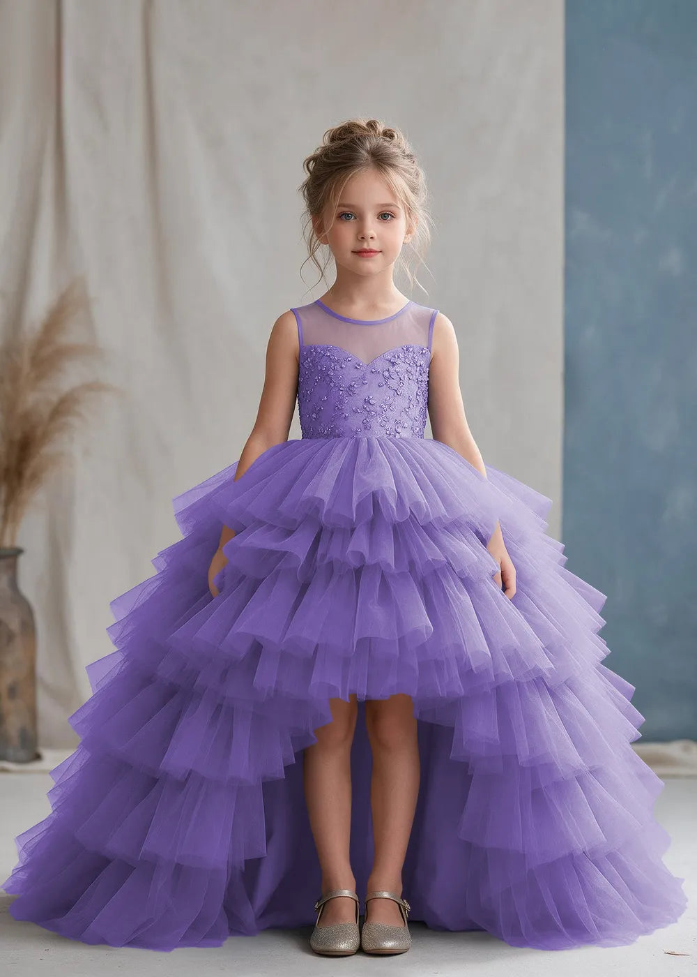 Front view of a child wearing a lavender flower girl dress with a sheer illusion neckline over a beaded bodice and a high-low tulle skirt with broad tiered ruffle layers.