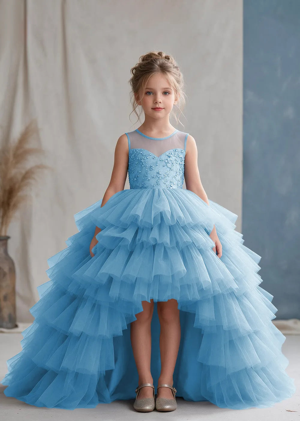 Front view of a child wearing a light blue flower girl dress with a sheer illusion neckline over a beaded bodice and a high-low tulle skirt with broad tiered ruffle layers.