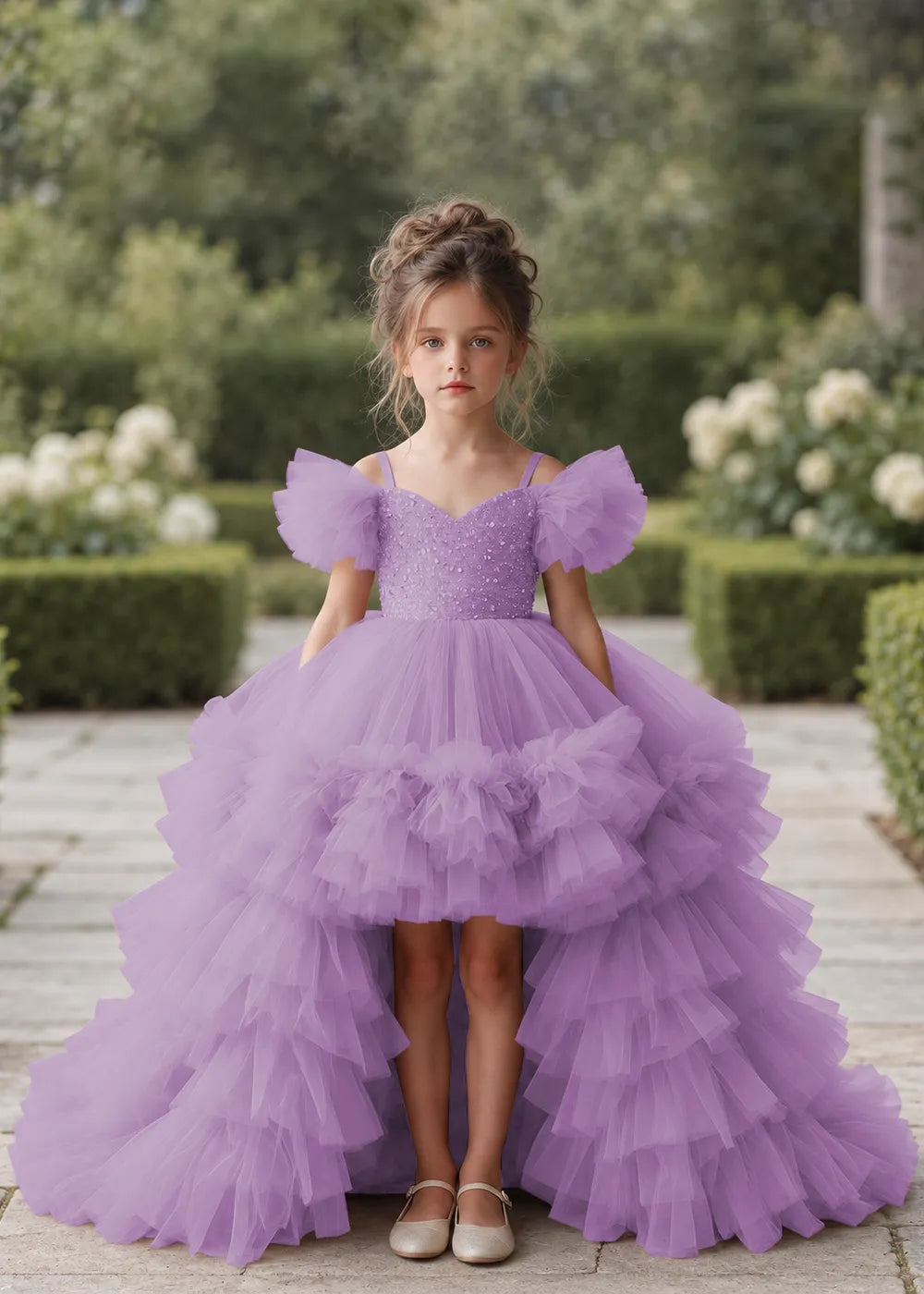 Front view of a child wearing a lilac flower girl dress with a V-neckline, a sequin-covered bodice and a tiered high-low tulle skirt with off-shoulder sleeve ruffles.
