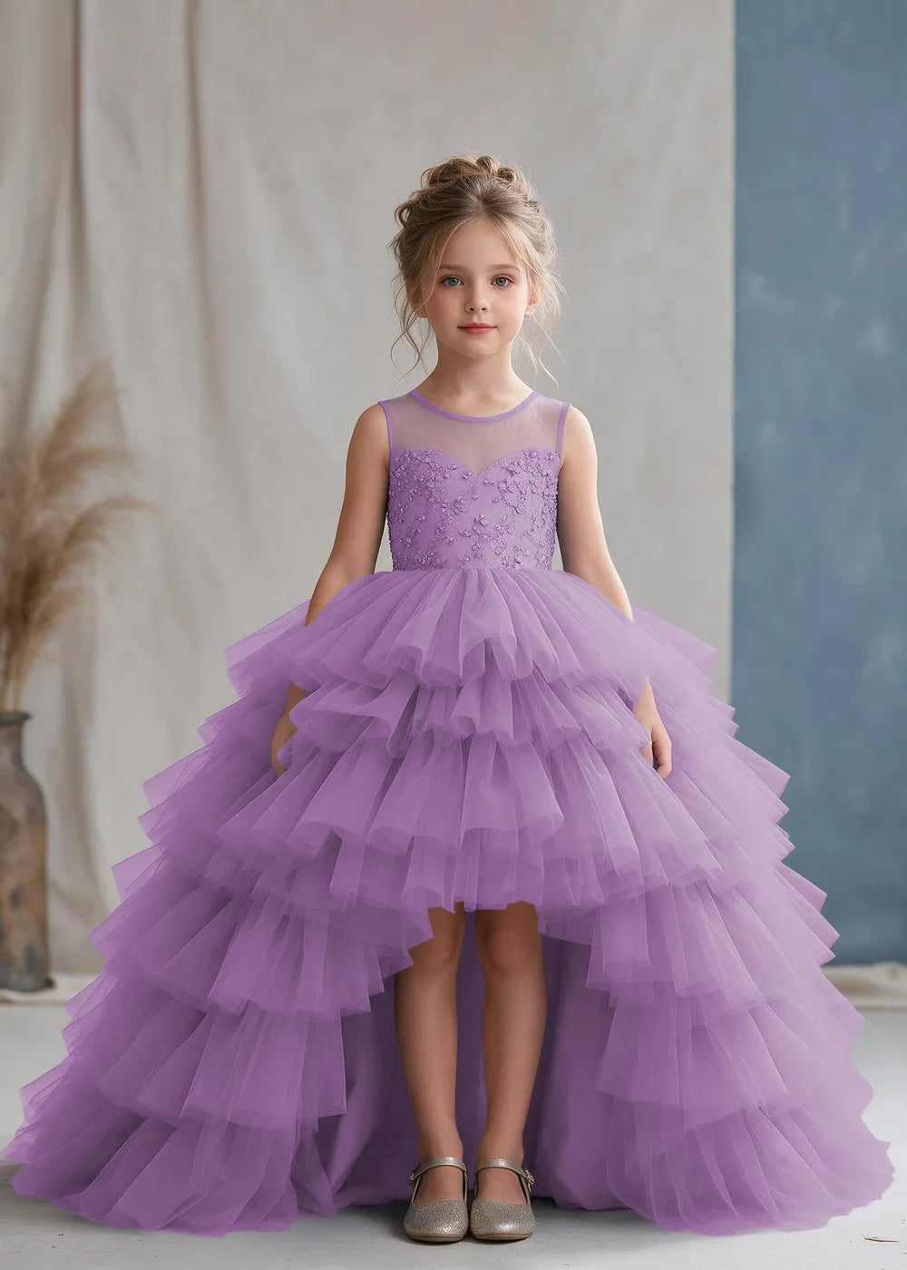 Front view of a child wearing a lilac flower girl dress with a sheer illusion neckline over a beaded bodice and a high-low tulle skirt with broad tiered ruffle layers.