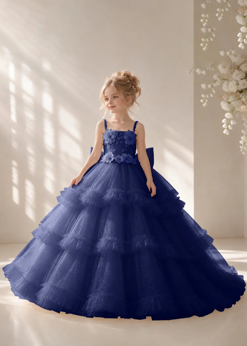 Front view of a child wearing a navy blue flower girl dress with beaded spaghetti straps, a bodice covered in three-dimensional fabric flowers and a multi-tiered tulle skirt.