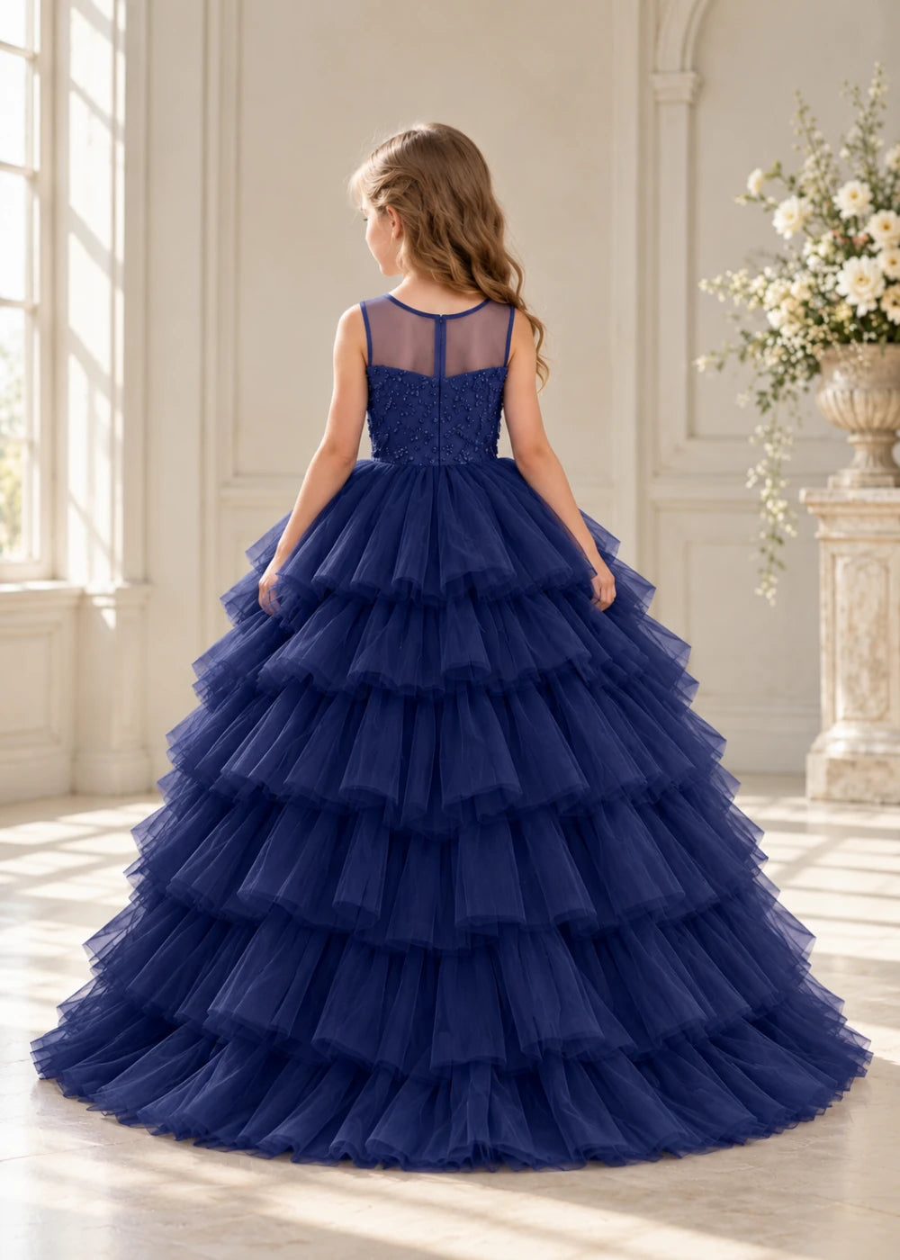 Rear view of a child wearing a navy blue flower girl dress with a sheer illusion neckline over a beaded bodice and a high-low tulle skirt with broad tiered ruffle layers.