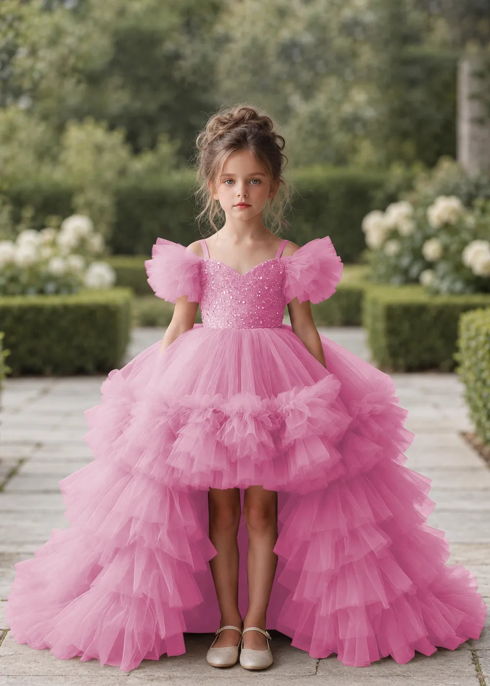 Front view of a child wearing a pearl pink flower girl dress with a V-neckline, a sequin-covered bodice and a tiered high-low tulle skirt with off-shoulder sleeve ruffles.