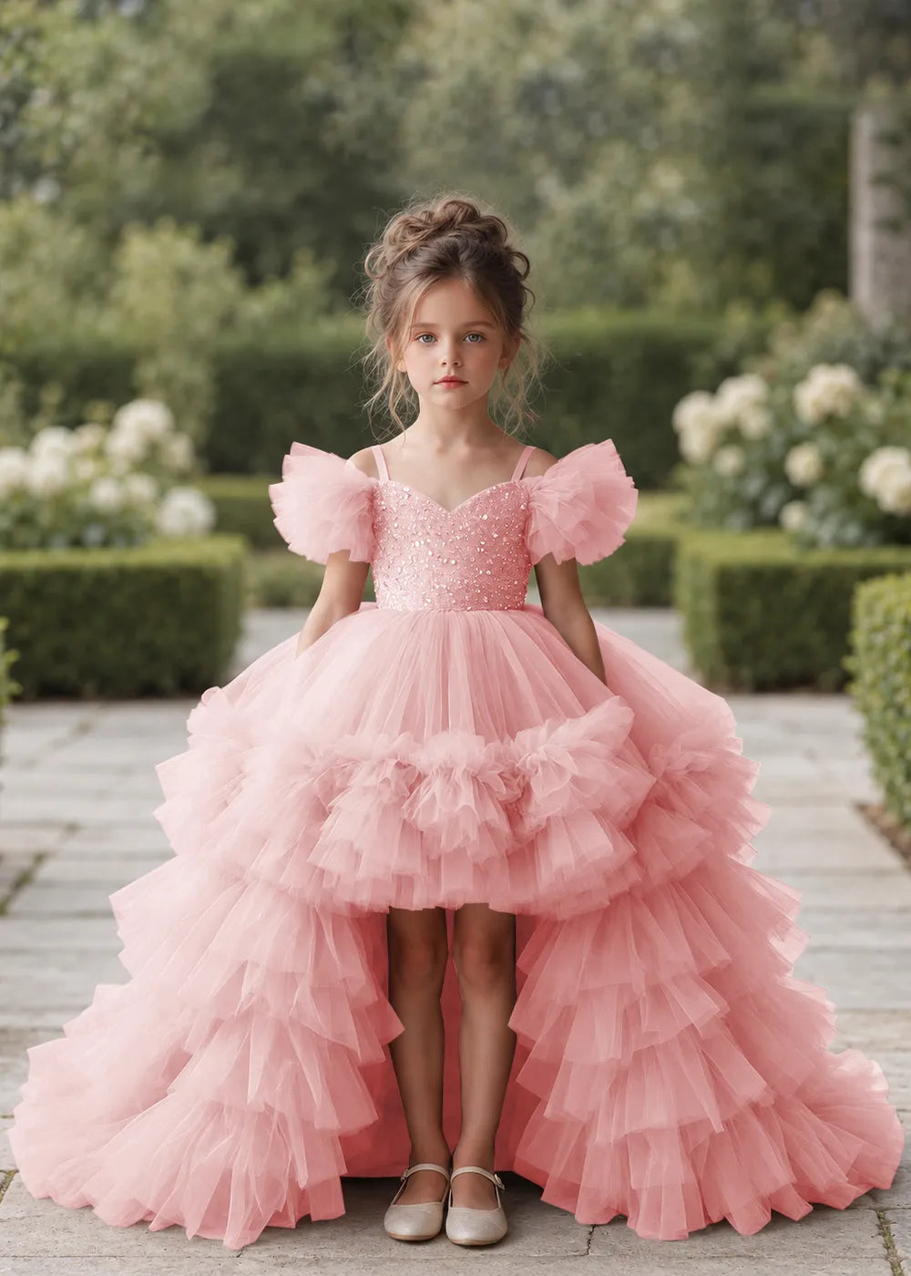 Front view of a child wearing a pink flower girl dress with a V-neckline, a sequin-covered bodice and a tiered high-low tulle skirt with off-shoulder sleeve ruffles.