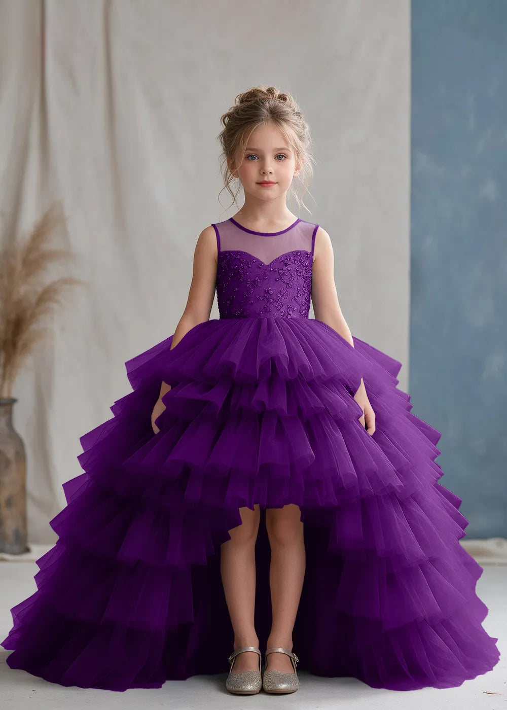 Front view of a child wearing a purple flower girl dress with a sheer illusion neckline over a beaded bodice and a high-low tulle skirt with broad tiered ruffle layers.