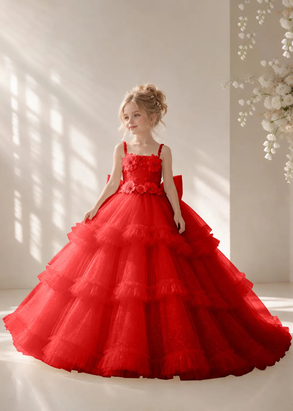 Front view of a child wearing a red flower girl dress with beaded spaghetti straps, a bodice covered in three-dimensional fabric flowers and a multi-tiered tulle skirt.