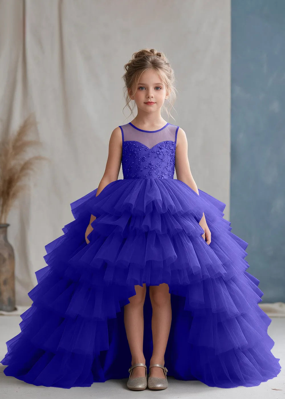 Front view of a child wearing a royal blue flower girl dress with a sheer illusion neckline over a beaded bodice and a high-low tulle skirt with broad tiered ruffle layers.