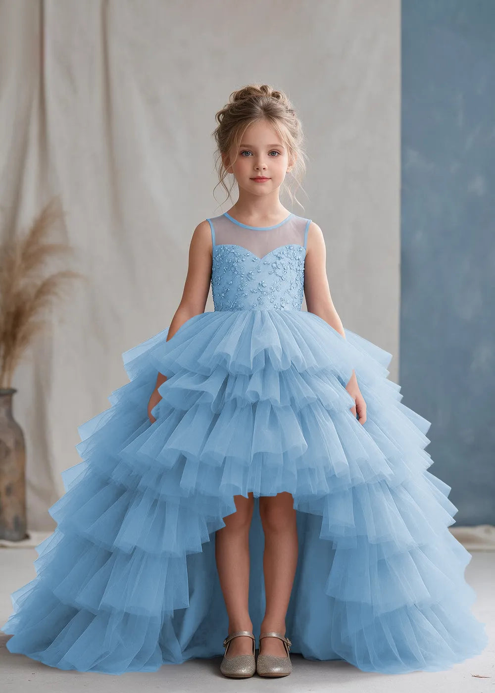 Front view of a child wearing a sky blue flower girl dress with a sheer illusion neckline over a beaded bodice and a high-low tulle skirt with broad tiered ruffle layers.