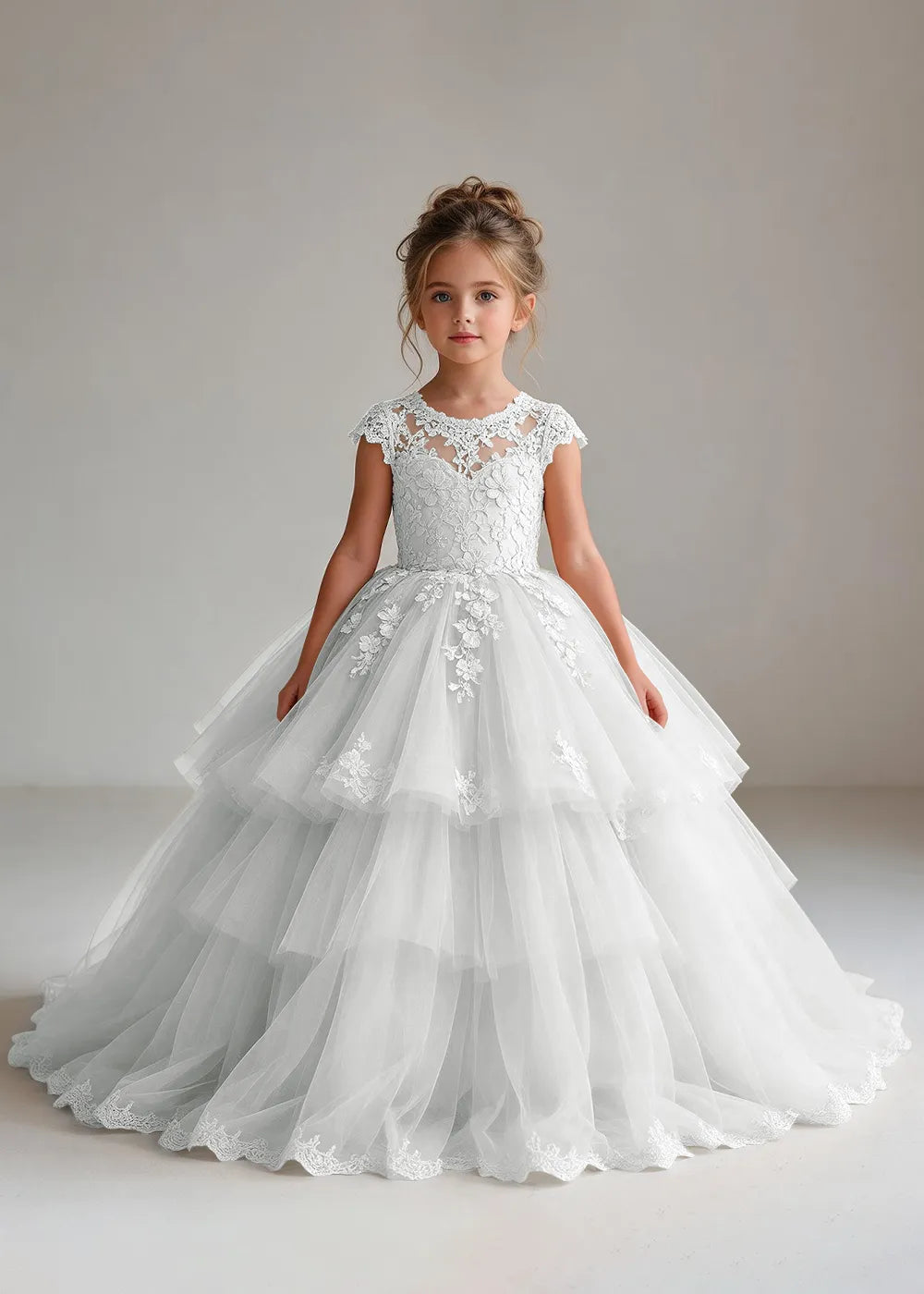 Front view of a child wearing a white flower girl dress with a sheer lace illusion neckline, cap sleeves and a tiered cake-style tulle skirt with a lace-trimmed hem.