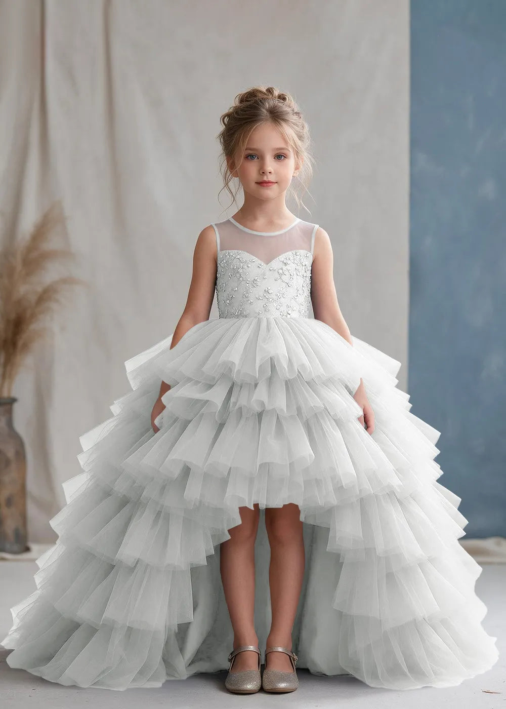 Front view of a child wearing a white flower girl dress with a sheer illusion neckline over a beaded bodice and a high-low tulle skirt with broad tiered ruffle layers.
