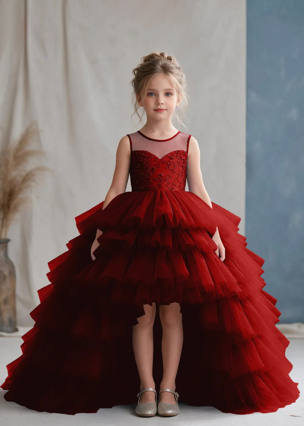 Front view of a child wearing a wine red flower girl dress with a sheer illusion neckline over a beaded bodice and a high-low tulle skirt with broad tiered ruffle layers.