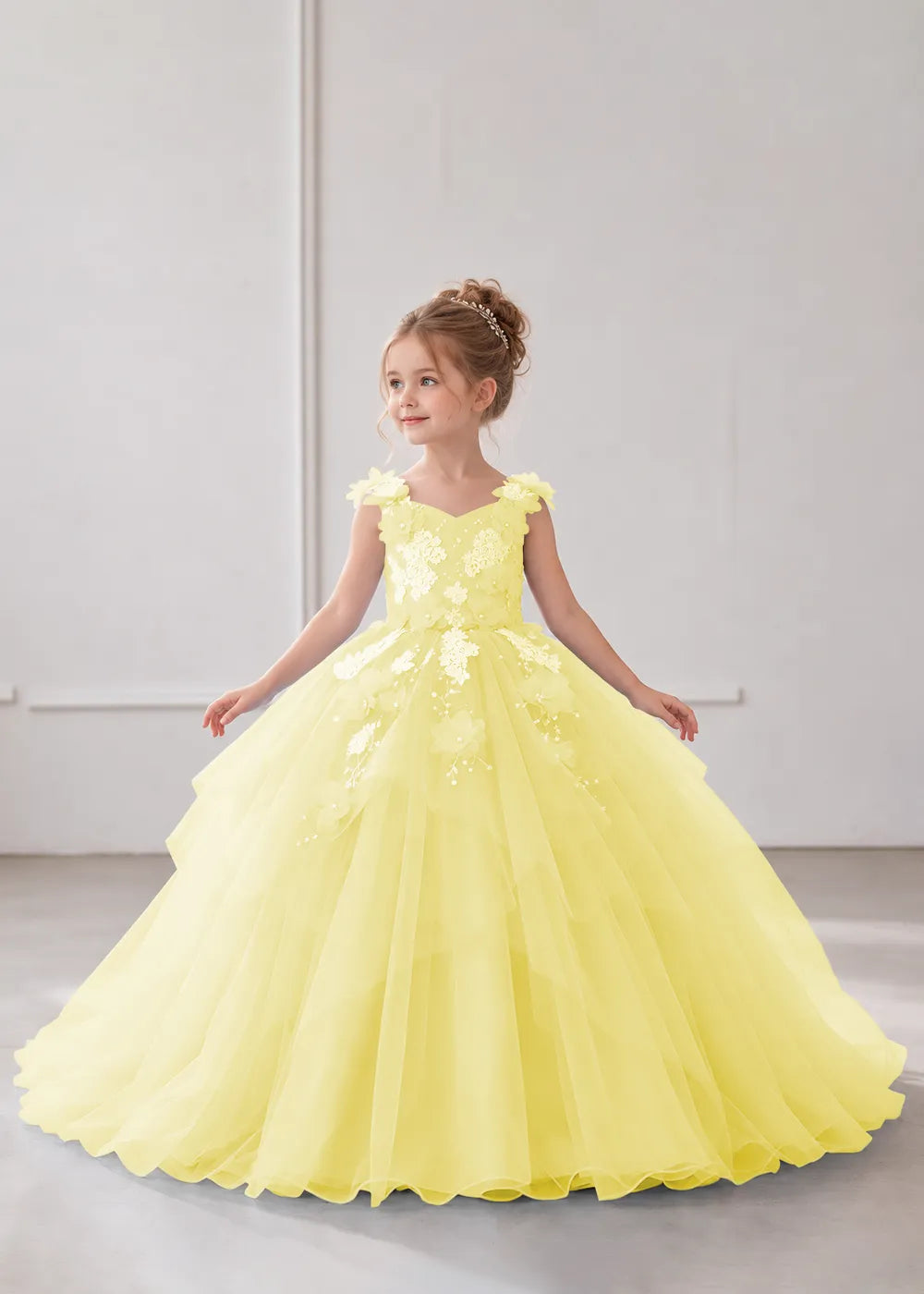 Front view of a child wearing a yellow flower girl dress with a V-neckline, floral shoulder appliques and lace and flower decoration across the bodice and hips.