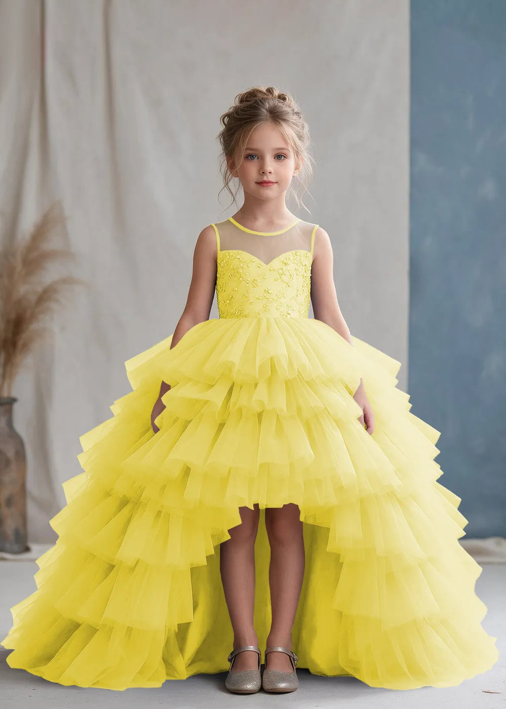 Front view of a child wearing a yellow flower girl dress with a sheer illusion neckline over a beaded bodice and a high-low tulle skirt with broad tiered ruffle layers.