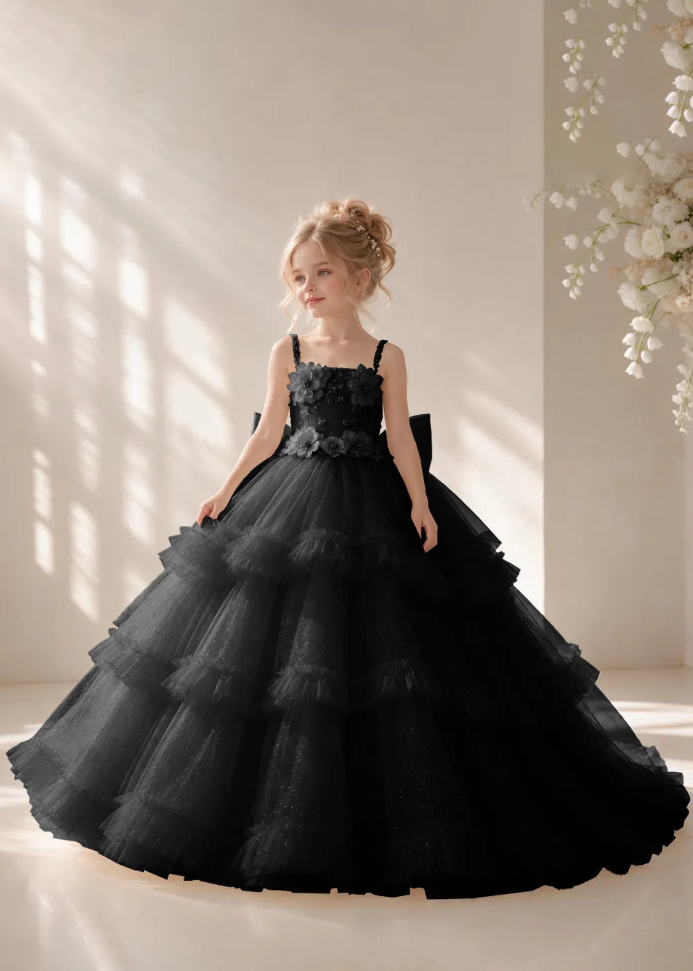 Front view of a child wearing a black flower girl dress with beaded spaghetti straps, a bodice covered in three-dimensional fabric flowers and a multi-tiered tulle skirt.
