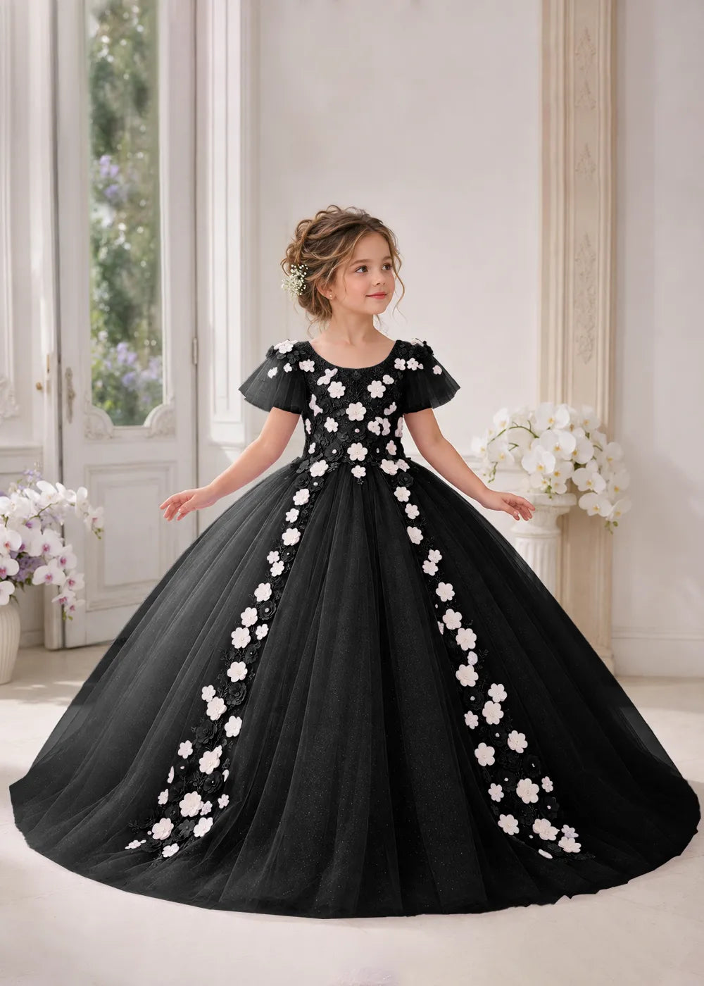 Front view of a child wearing a black flower girl dress with a round neckline, sheer flutter cap sleeves and a floral vine cascading down both sides of a full tulle skirt.