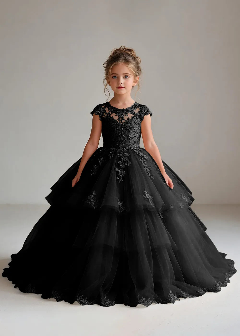 Front view of a child wearing a black flower girl dress with a sheer lace illusion neckline, cap sleeves and a tiered cake-style tulle skirt with a lace-trimmed hem.