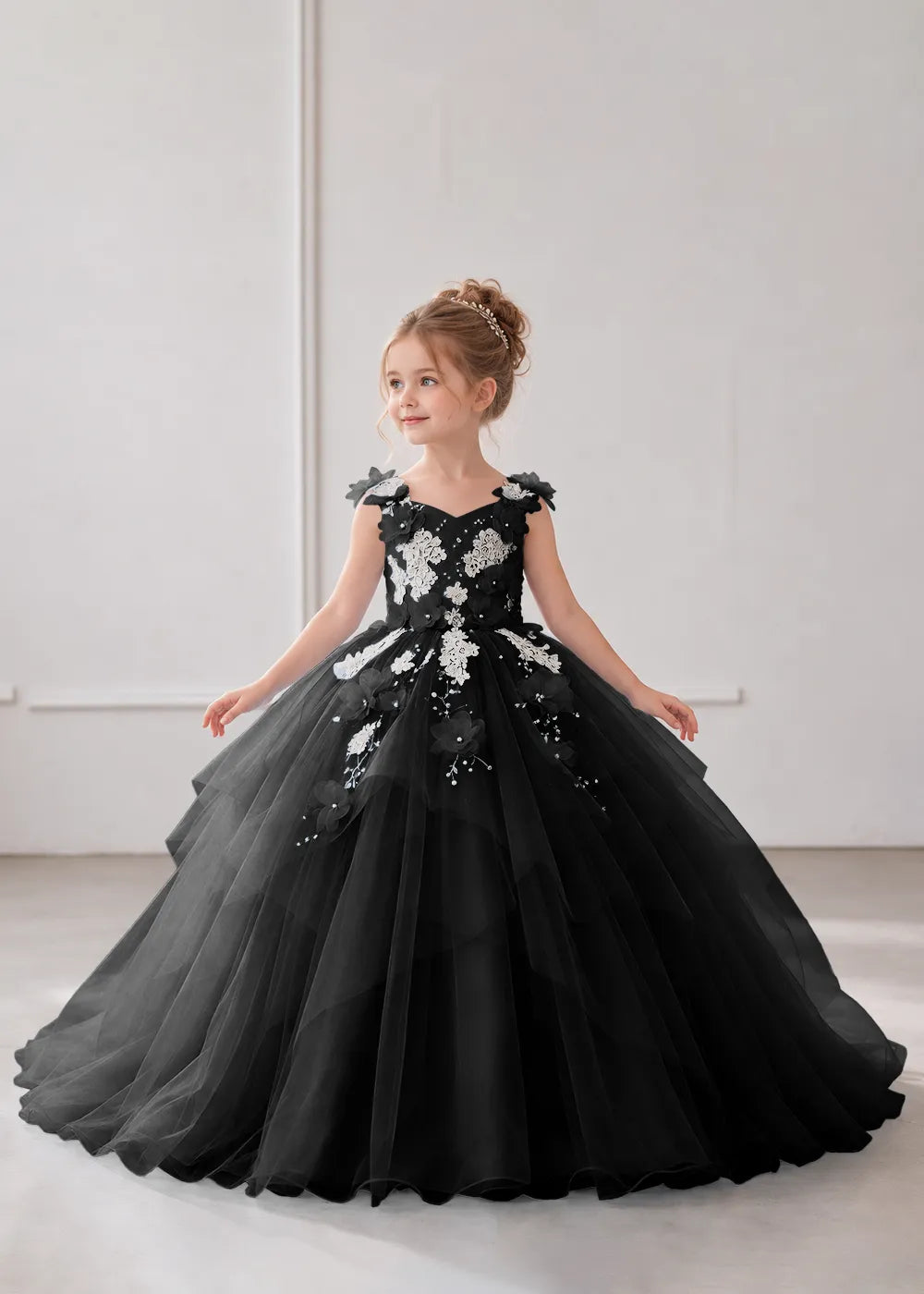 Front view of a child wearing a black flower girl dress with a V-neckline, floral shoulder appliques and lace and flower decoration across the bodice and hips.