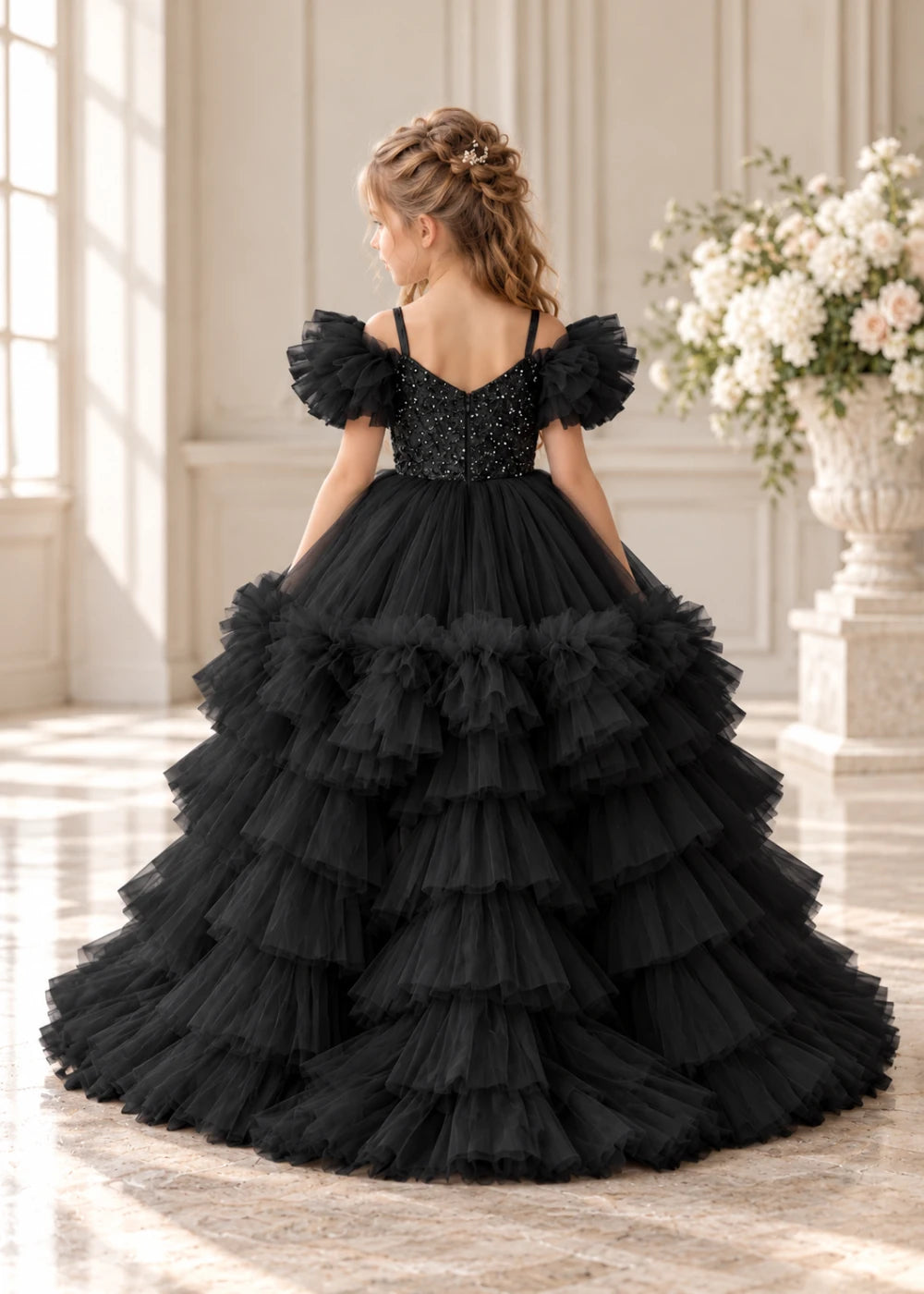 Rear view of a child wearing a black flower girl dress with a V-neckline, a sequin-covered bodice and a tiered high-low tulle skirt with off-shoulder sleeve ruffles.