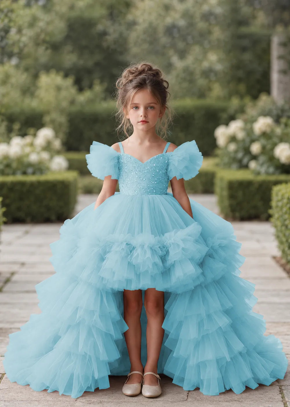 Front view of a child wearing a blue flower girl dress with a V-neckline, a sequin-covered bodice and a tiered high-low tulle skirt with off-shoulder sleeve ruffles.