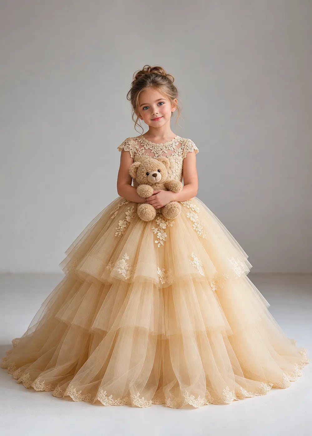 The front view of a child wearing a Champagne flower girl dress with a sheer lace illusion neckline, cap sleeves and a tiered cake-style tulle skirt with a lace-trimmed hem.