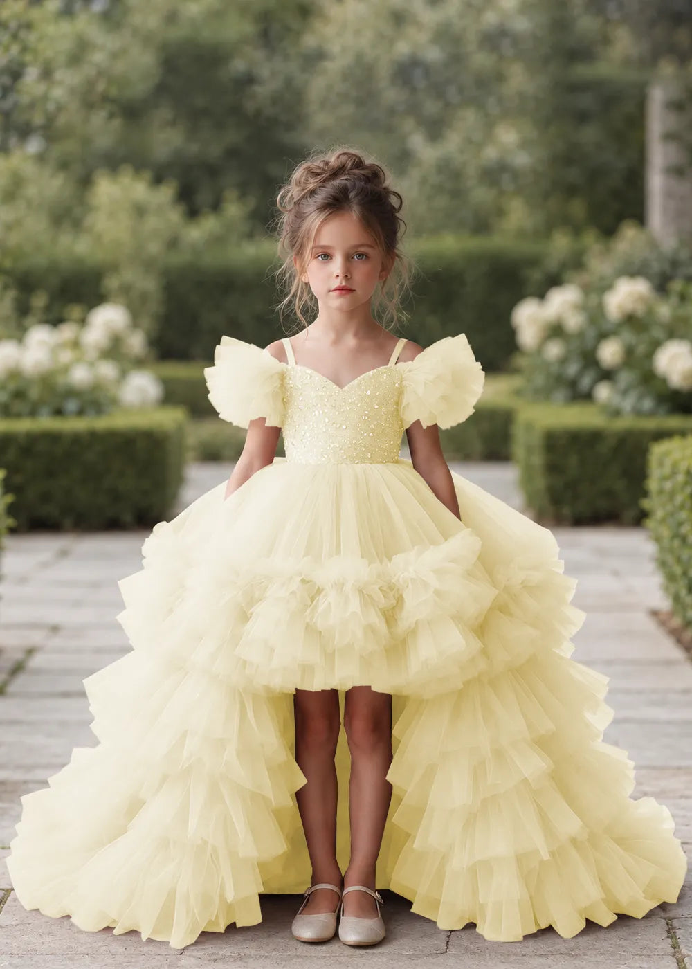 Front view of a child wearing a champagne flower girl dress with a V-neckline, a sequin-covered bodice and a tiered high-low tulle skirt with off-shoulder sleeve ruffles.