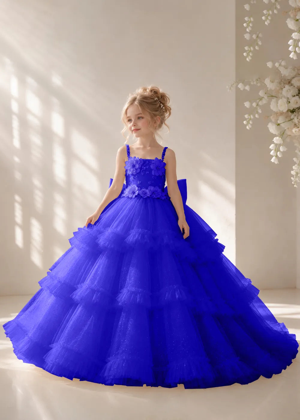Front view of a child wearing a deep royal blue flower girl dress with beaded spaghetti straps, a bodice covered in three-dimensional fabric flowers and a multi-tiered tulle skirt.