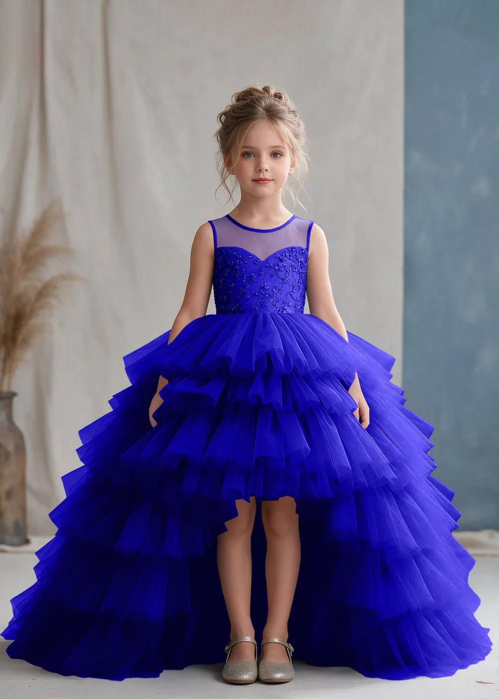 Front view of a child wearing a deep royal blue flower girl dress with a sheer illusion neckline over a beaded bodice and a high-low tulle skirt with broad tiered ruffle layers.